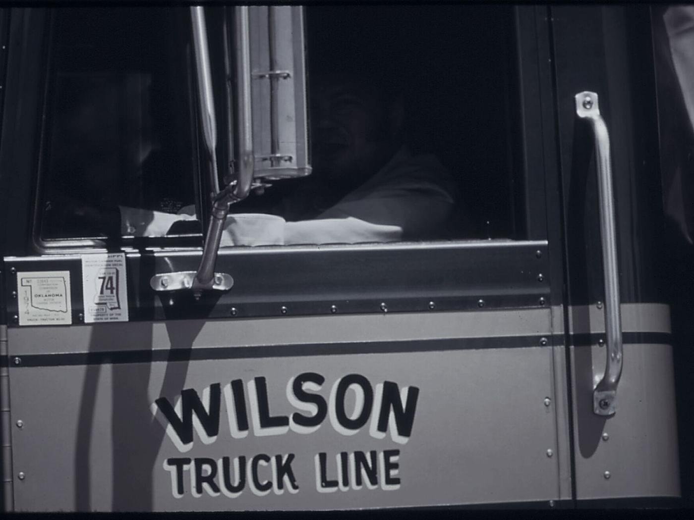 A driver at the wheel of a truck cab, seen through the open window.